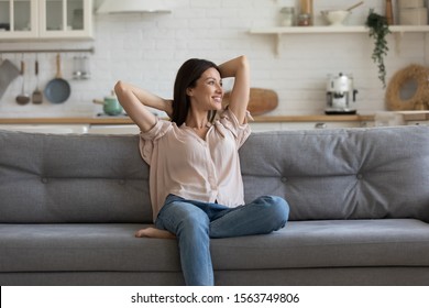 Smiling Positive 30s Woman Sitting On Cozy Couch, Crossing Hands Behind Head, Resting At Home. Young Peaceful Lady Looking Away, Planning Day Or Future, Enjoying Lazy Weekend Alone In Own Apartment.