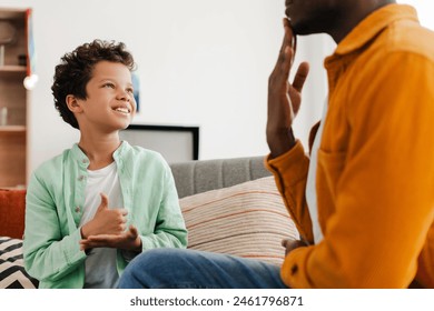 Smiling, positive African American little boy and his dad communicating with sign language while sitting on comfortable sofa in cozy home. Concept of health care, relationships - Powered by Shutterstock