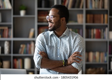 Smiling positive African American young man wearing glasses dreaming about good future and visualizing, new opportunities, looking to aside, standing with arms crossed in modern cabinet - Powered by Shutterstock