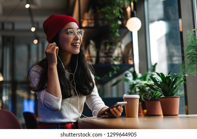 Smiling, Positive Asian Woman With Beautiful Emotional Face Wearing Eyeglass Listening To Music In Coffee Shop. Pretty Hipster Female Holding Mobile Phone, Laughing, Dreaming, Drinking Coffee. 