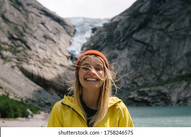 Smiling, positive blonde woman with wild hair in full face on background of blue ice tongue of Briksdal glacier that slides from the giant rocky mountain and melts into cold lake in Norway - Powered by Shutterstock