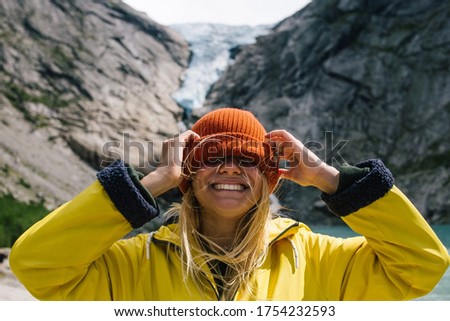 Smiling, positive blonde woman with wild hair puts a hat on her face on background of blue ice tongue of Briksdal glacier that slides from the giant rocky mountain and melts into cold lake in Norway