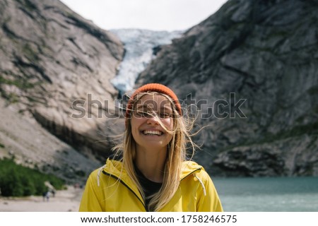 Smiling, positive blonde woman with wild hair in full face on background of blue ice tongue of Briksdal glacier that slides from the giant rocky mountain and melts into cold lake in Norway