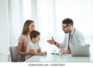 Smiling Positive Gp Pediatrician In Eyeglasses And Medical Uniform Stroking Shoulder Of Little Boy Patient, Encouraging At Clinic Checkup. Young Mother Holding Small Kid Son On Laps, Greeting Doctor.