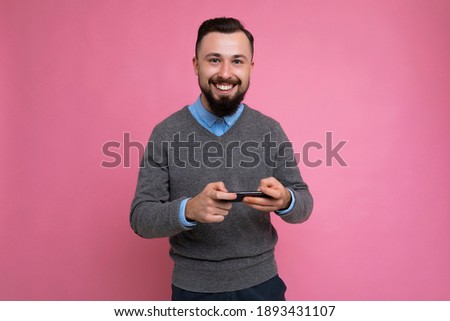 Smiling positive handsome good looking brunet bearded young man wearing grey sweater and blue shirt isolated on pink background with empty space holding in hand and using mobile phone communicating