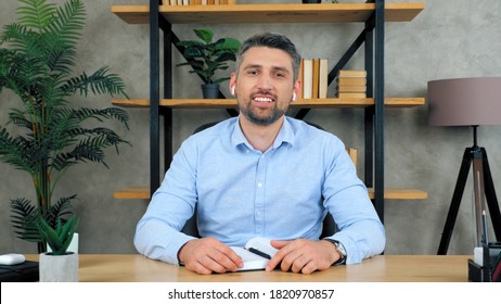 Smiling Positive Happy Business Man Sitting On Chair At Table In Home Office Wearing Wireless Earphones Looks Into Camera Listening To Interlocutor On Video Call Chat Conference, Web Cam Concept