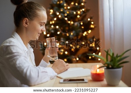 Smiling positive happy woman writing in a notebook Christmas bucket list on table with flower and candle, posing in living room with xmas tree, drinking water from glass.
