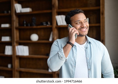 Smiling and positive indian guy in casual shirt talking by the smartphone in the modern office. Cheerful mixed-race man has phone conversation. Joyful male employee speaking on mobile phone indoors - Powered by Shutterstock