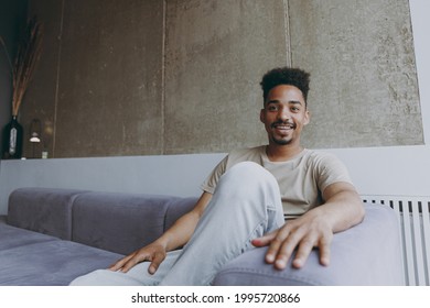 Smiling Positive Lazy Fun Young African American Man In Beige T-shirt Sweatpants Sitring On Fashionable Grey Sofa Indoors Apartment Near Heating Battery Procrastinate, Rest On Weekends Stay At Home.