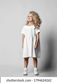 Smiling positive little blonde curly kid girl in white casual dress and sneakers is looking at copy space at upper corner. Stylish comfortable everyday fashion for children concept