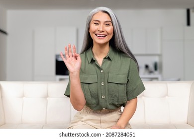 Smiling And Positive Middle-aged Asian Woman Holding Video Call, Looking And Waving Into Camera, Mid-age Multiracial Gray-haired Tutor Holding Virtual Meeting, Involved Webinar, Webcam View