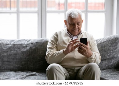 Smiling Positive Old Man Sit On Couch In Living Room Holding Mobile Phone Enjoy Usage Feels Satisfied Using Messenger, Makes Call To Friend Or Grownup Children, Spend Time Web Surfing On-line Concept