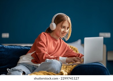 Smiling positive teen girl in headphones watching video movie on laptop sitting on couch at home. Leisure, recreation, entertainment pastime of teenager choosing film, cartoon to watch on computer.  - Powered by Shutterstock