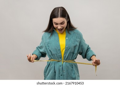 Smiling positive woman holding tape line on her waist, being amazed with her body parameters after slimming, wearing casual style jacket. Indoor studio shot isolated on gray background.