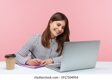 Smiling positive woman office worker in striped shirt making notes with pencil at notepad looking at laptop display, has video call conference. Indoor studio shot isolated on pink background