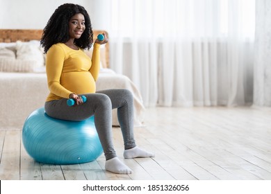 Smiling pregnant black lady exercising with barbells while sitting on fitness ball, looking at camera, copy space, bedroom interior. African american expecting lady training at home, using yoga ball - Powered by Shutterstock