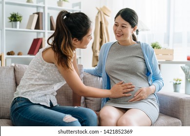 smiling pregnant woman communicates with girl friend indoors. young future motherhood lady showing her belly with baby expect bestie hands touching feeling. two female sitting on sofa chatting home - Powered by Shutterstock