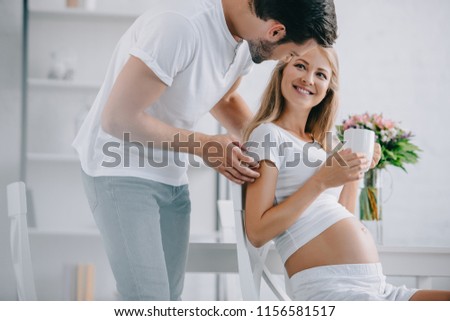 smiling pregnant woman with cup of tea resting on chair with husband near by at home