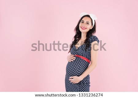 Smiling pregnant woman in dotted dress touching belly isolated on pink