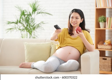 Smiling pregnant woman eating apple and watching tv at home