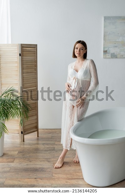 Smiling pregnant woman in robe looking at camera\
near bathtub in\
bathroom