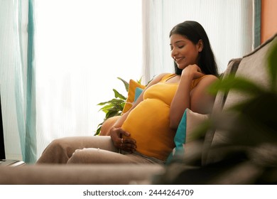 Smiling pregnant woman sitting on sofa at home with holding her abdomen - Powered by Shutterstock