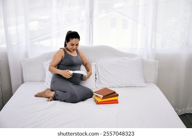 Smiling pregnant woman sitting on a bed, using headphones on her belly, with books nearby, promoting relaxation and bonding - Powered by Shutterstock