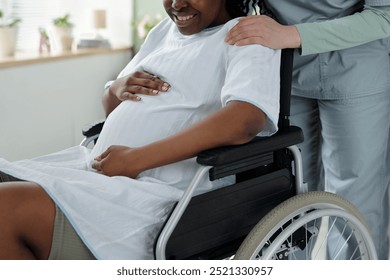 Smiling pregnant woman sitting in wheelchair holding belly while nurse stands beside in medical setting indicating support and care for expectant mothers - Powered by Shutterstock