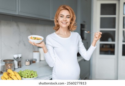 A smiling pregnant woman stands in a modern kitchen, holding a bowl of salad in one hand and a spoon in the other. Fresh fruits and vegetables are on the counter nearby. - Powered by Shutterstock