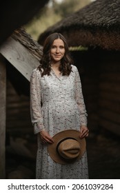 A Smiling Pregnant Woman Is Staying Near Wooden Barns With A Cane Roof In An Eastern European Village. A Happy Pregnancy On The Farm. A Lady In A Long Dress And A Hat In The Countryside In The Evening