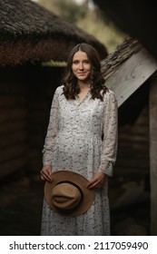 A Smiling Pregnant Woman Is Staying Near Wooden Barns With A Cane Roof In An Eastern European Village. A Happy Pregnancy On The Farm. A Lady In A Long Dress And A Hat In The Countryside In The Evening