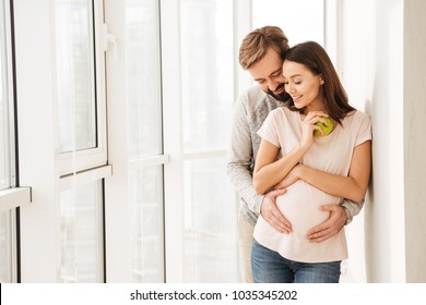 Smiling pregnant young couple hugging while standing together at a window