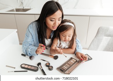 Smiling Pretty Asian Woman Doing Make Up While Sitting With Her Little Daughter At The Kitchen