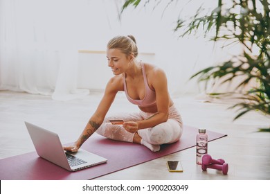 Smiling pretty beautiful young strong sporty fitness woman wearing pink tracksuit doing yoga exercises using laptop computer hold credit bank card sitting and stretching on mat floor at home indoor - Powered by Shutterstock