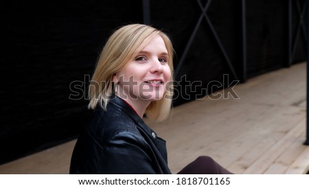 Smiling pretty blond young woman in black leather jacket and red shirt sitting outdoor on the street in the city. Spending time walking and having rest concept