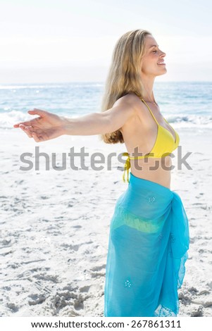 Smiling pretty blonde standing by the sea arms outstretched at the beach