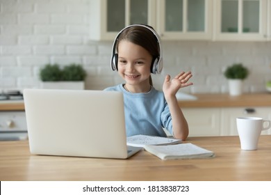 Smiling pretty cute little 7 years old kid girl in headphones looking at laptop screen, waving hello gesture. Happy adorable caucasian child starting distant learning lesson with teacher at home. - Shutterstock ID 1813838723
