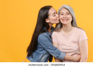 Smiling Pretty Family Asian Women Girls Gray-haired Mother And Brunette Daughter In Casual Clothes Posing Hugging Kissing In Cheek Looking Camera Isolated On Yellow Color Background Studio Portrait