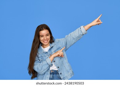 Smiling pretty freckled gen z brunette Latin girl, cute cheerful happy Hispanic teen student wearing denim outfit looking at camera pointing aside at copy space standing isolated on blue background. - Powered by Shutterstock