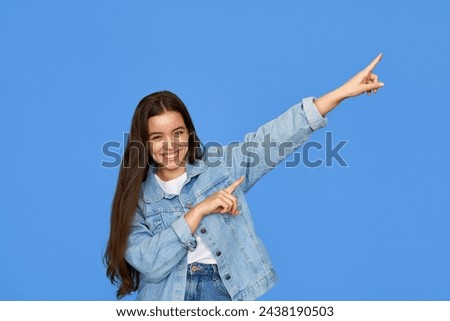 Smiling pretty freckled gen z brunette Latin girl, cute cheerful happy Hispanic teen student wearing denim outfit looking at camera pointing aside at copy space standing isolated on blue background.
