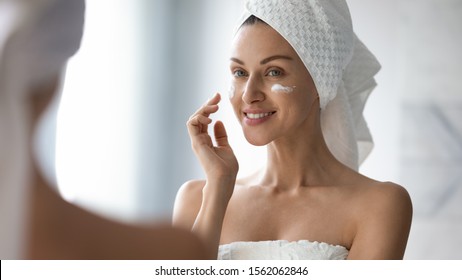 Smiling Pretty Lady Put Anti-ageing Hydrating Moisturizing Lifting Nourishing Facial Cream In Bathroom, Happy Young Woman Wrap In Towel Look In Mirror Apply Creme On Face Skin Care Treatment Concept