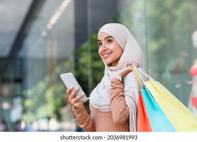 Smiling pretty millennial arabian lady in hijab enjoys online shopping with bags, surfing on phone near mall and look at empty space. New mobile app for messages from store about discount and sale - Powered by Shutterstock