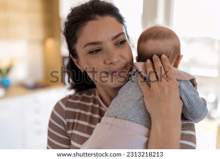 Smiling pretty mum holding infant on shoulder and enjoying tender moment in hug at home beside window. Mothernity love, care and joy concept