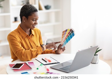 Smiling Pretty Short-haired Stylish Millennial Black Woman Working As Web Designer, Sitting At Workdesk In Front Of Laptop, Holding Color Palette, Making Website, White Office Interior, Copy Space