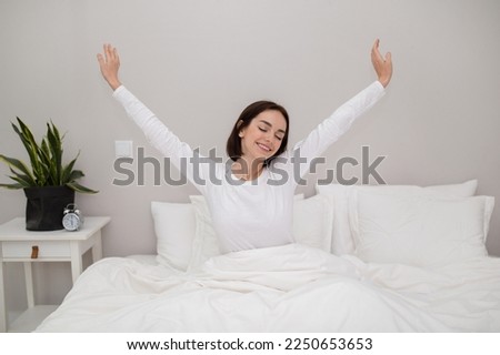 Smiling pretty slim brunette young woman enjoying happy morning, sitting in comfy bed in white cozy bedroom with plant and alarm on bed side table, stretching with closed eyes