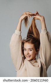 Smiling pretty teenage girl with brackets pulling her hair up, playing cutie. Looking at the camera. Against grey background. In a warm beige longsleeve.