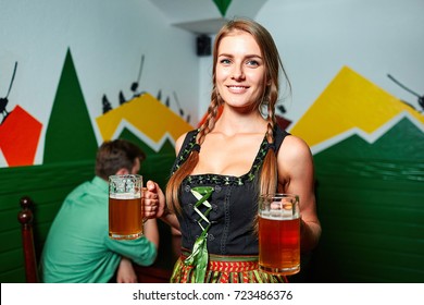 Smiling Pretty Waitress In German Octoberfest Suit Posing With Two Beer Glasses. Happy Couple On The Background.