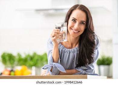 Smiling Pretty Woman Holds A Glass Of Water Leaning On Kitchen Desk.