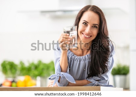 Smiling pretty woman holds a glass of water leaning on kitchen desk.