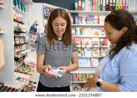 Smiling pretty woman seller shows a palette of vibrant watercolor paints, to pregnant woman artist, a customer in a school stationery and art store. Painting. Fine art. Hobby, leisure, people, retail
